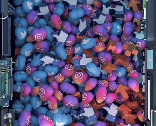 A top-down view of a vibrant pile of colorful icons and symbols representing various social media platforms. Icons include blue Twitter birds, Instagram logos, Facebook-like thumbs up, and orange up arrows. The symbols are situated inside a futuristic, mechanical enclosure, possibly resembling a server or machine.