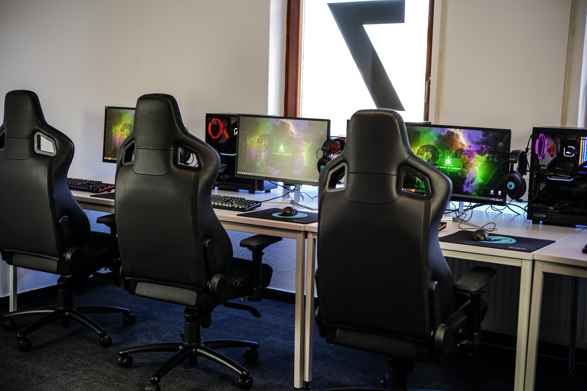 A row of gaming stations equipped with large monitors, gaming chairs, keyboards, and headphones. The setup is organized and ready for gaming, featuring a colorful screen display. The room has a modern and tech-savvy ambiance.