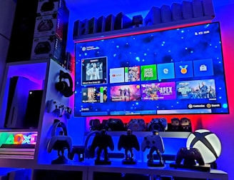 A modern gaming setup featuring a large wall-mounted screen displaying a gaming console interface. The room is decorated with gaming-themed items such as controllers and collectibles. The ambient lighting includes shades of blue and red, creating a vibrant atmosphere.