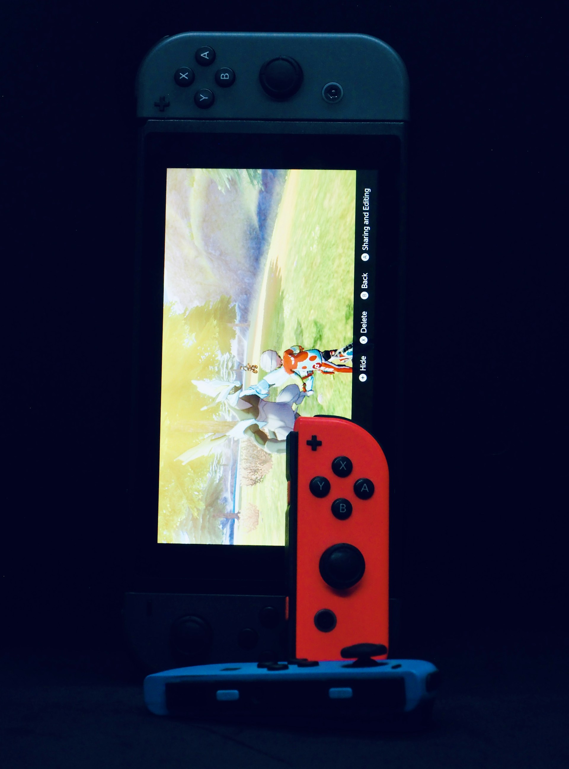 A gaming console with a screen displaying a video game scene, accompanied by two controllers, one colored red and the other blue. The devices are set against a dark backdrop, emphasizing the vibrant colors of the controllers.