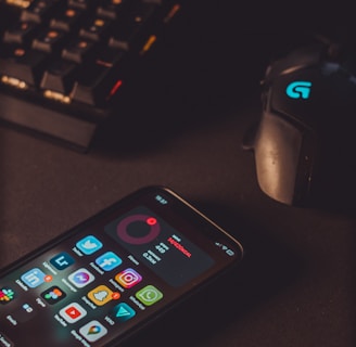 A smartphone displaying various social media app icons is placed on a dark surface. Nearby, a gaming mouse with a glowing logo and a mechanical keyboard with illuminated keys are visible. The lighting is low and focused, creating a moody atmosphere.