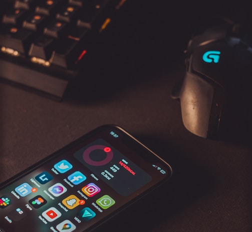 A smartphone displaying various social media app icons is placed on a dark surface. Nearby, a gaming mouse with a glowing logo and a mechanical keyboard with illuminated keys are visible. The lighting is low and focused, creating a moody atmosphere.
