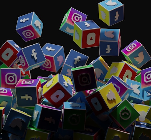 A collection of colorful 3D cubes displaying logos of various social media platforms, including Facebook, Instagram, Twitter, YouTube, and others. The cubes appear to be falling and tumbling over each other against a dark background.